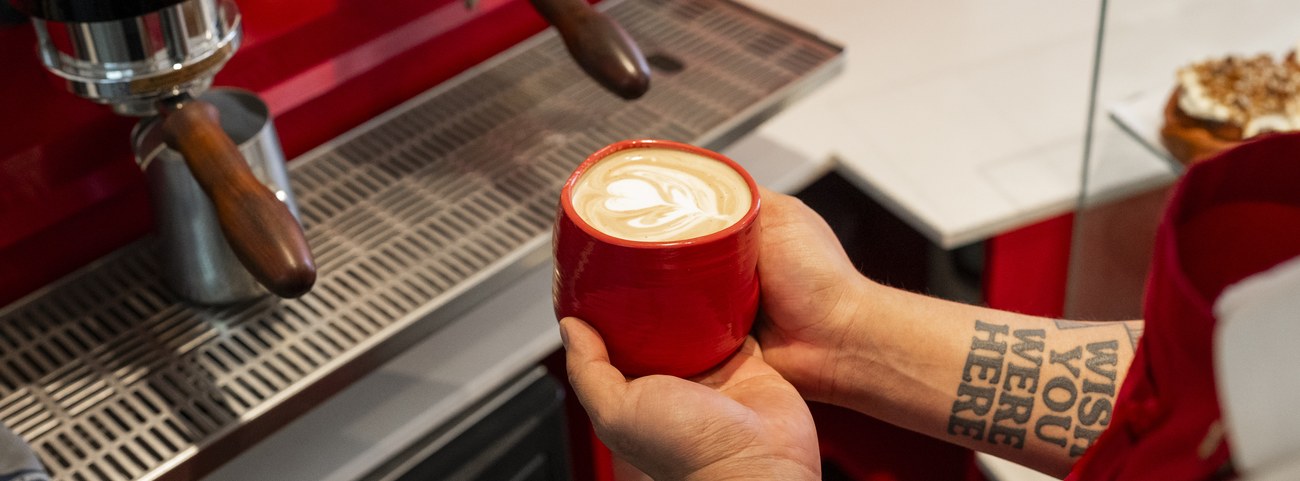 Manos del barista sosteniendo una taza roja con arte latte junto a la máquina