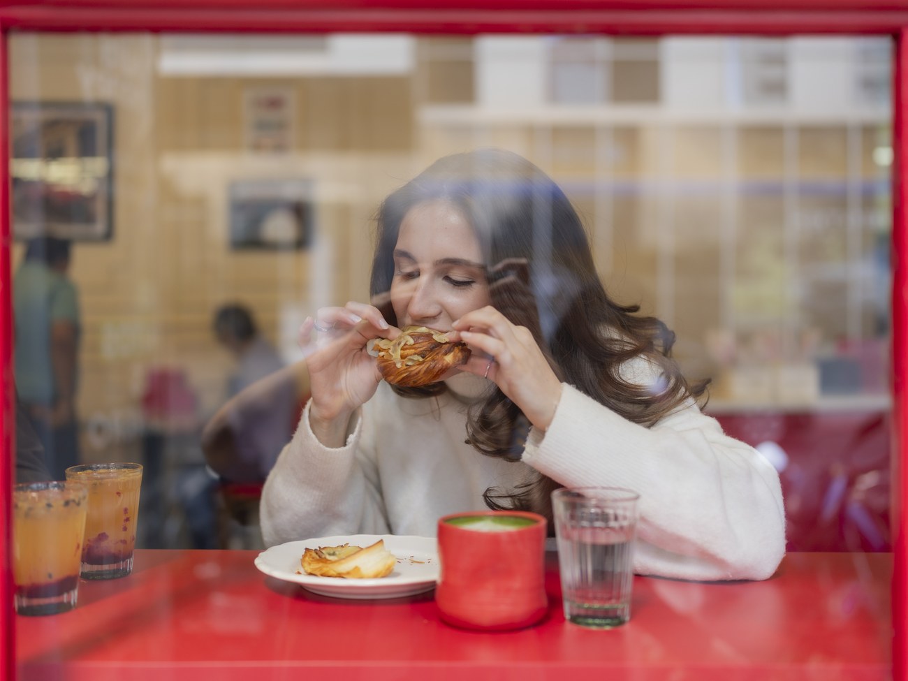 Clienta comiendo una pieza de pan, vista desde la calle a través del marco rojo de la ventana